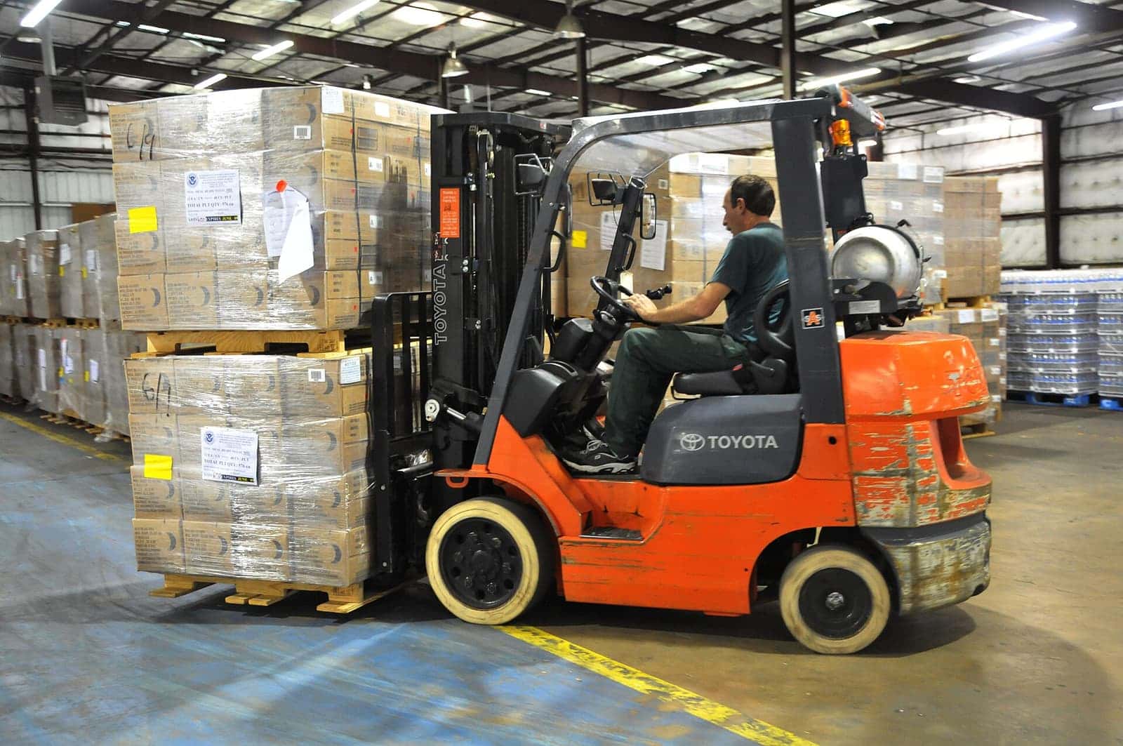 Operator mengemudikan forklift oranye yang membawa tumpukan kardus di dalam gudang