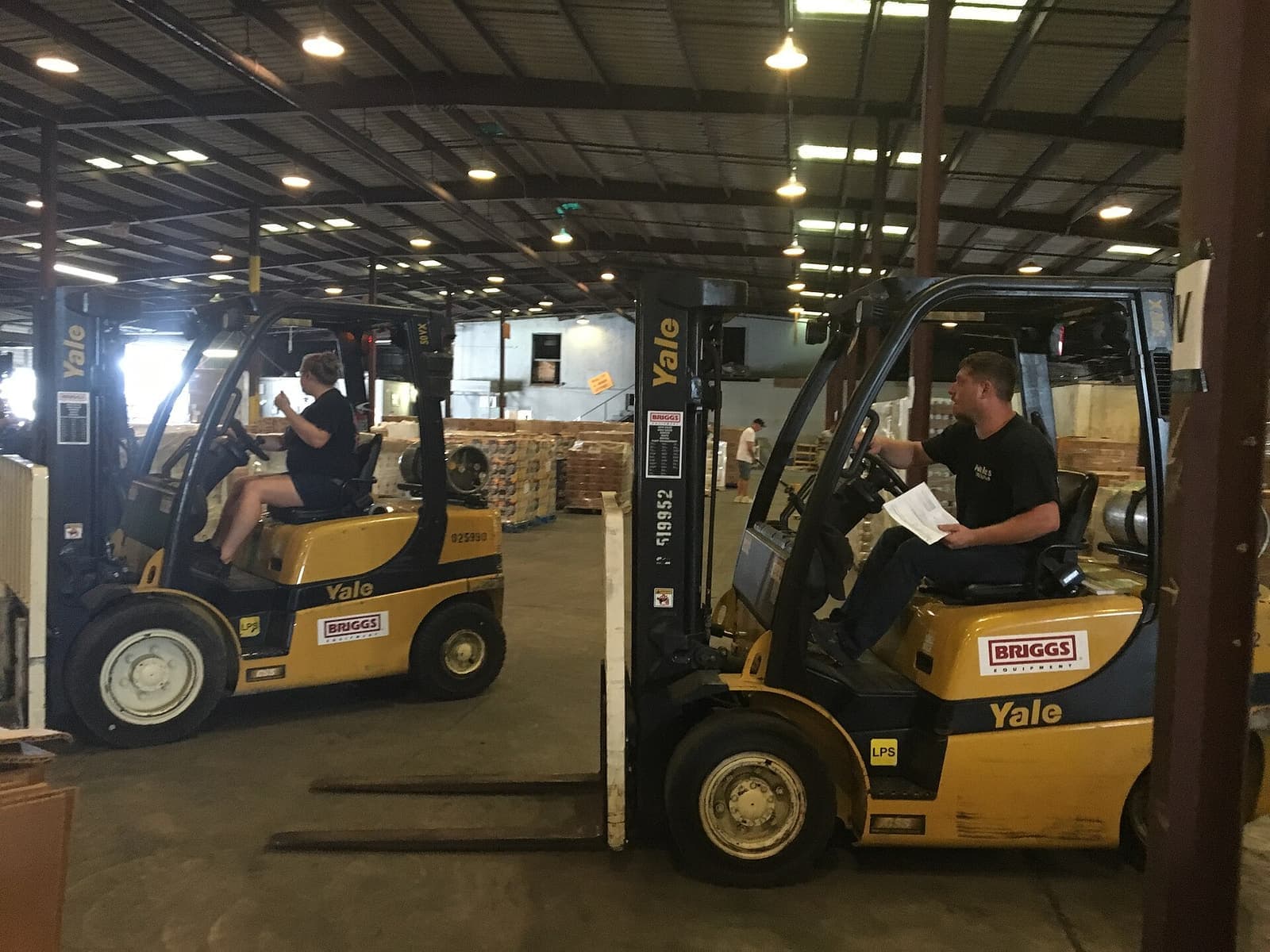 Dua operator duduk di atas forklift kuning di area gudang